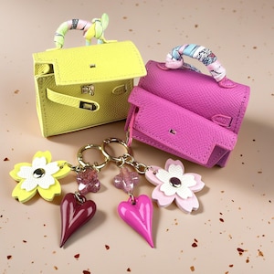 May include: Two miniature handbags in bright yellow and vibrant pink, each featuring a textured finish and a handle wrapped in a colourful patterned ribbon. Attached to each bag is a gold-toned keychain with a flower charm and a glossy heart pendant.