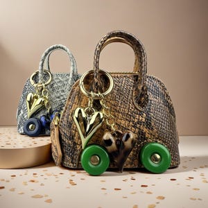 Könnte beinhalten: Zwei kuppelförmige Miniaturhandtaschen mit Schlangenledermuster. Die vordere Tasche ist braun und schwarz mit zwei grünen Rädern und einem goldenen Herzanhänger. Die hintere Tasche ist grau und schwarz mit zwei dunkelblauen Rädern und einem Herz.