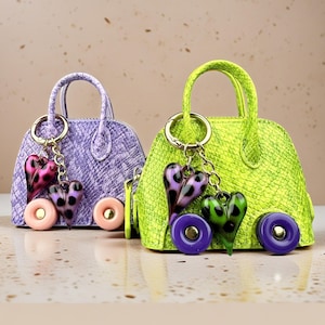 May include: Two miniature handbags with a textured snakeskin pattern, one in lavender and one in lime green. Each bag features two round wheels at the base, a top handle, and a gold-toned keychain with two dangling heart-shaped charms.