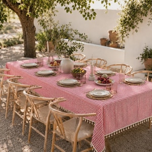 Puede incluir: Una mesa de comedor rectangular cubierta con un mantel de cuadros vichy rojos y blancos con un borde de flecos blancos. La mesa está dispuesta con platos blancos, manteles individuales trenzados, cristalería y un jarrón central.