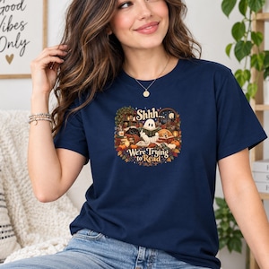 Può includere: Una t-shirt a maniche corte blu navy con una stampa grafica colorata raffigurante un fantasma, un gatto e un cane che leggono libri circondati da foglie autunnali, zucche e lanterne, con il testo "Shhh... We're Trying to Read" al centro.