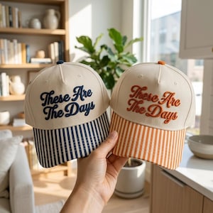 May include: Two baseball caps with off-white crowns and embroidered text reading These Are The Days. One cap features a navy blue striped brim and navy blue text, while the other has an orange striped brim and matching orange text.