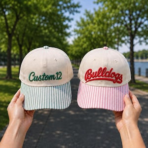 May include: Two baseball caps with off-white crowns. The left cap features green embroidered text reading Custom 12 and a green and white striped brim. The right cap features red embroidered text reading Bulldogs and a pink and white striped brim.