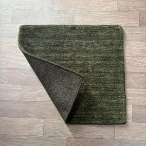 May include: A rectangular olive green rug with a textured, fibrous surface and a folded corner revealing a dark, woven backing. The rug features a consistent, dense pile and a uniform color throughout, showcasing the contrast between the top and bottom.