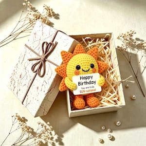 Può includere: Un peluche a forma di sole lavorato all'uncinetto in giallo e arancione con un viso sorridente, che tiene un cartoncino rettangolare bianco con la scritta Happy Birthday You are truly SUN-sational, in una scatola regalo bianca.