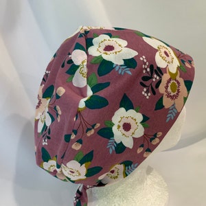 May include: A mauve scrub hat with a white and green floral print. The hat has a tie at the back.