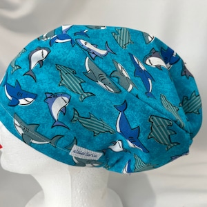 May include: A blue surgical scrub cap featuring a pattern of various cartoon sharks in shades of blue, gray, and white. The cap is made of fabric and has a textured background. The cap is designed to cover the head and hair.
