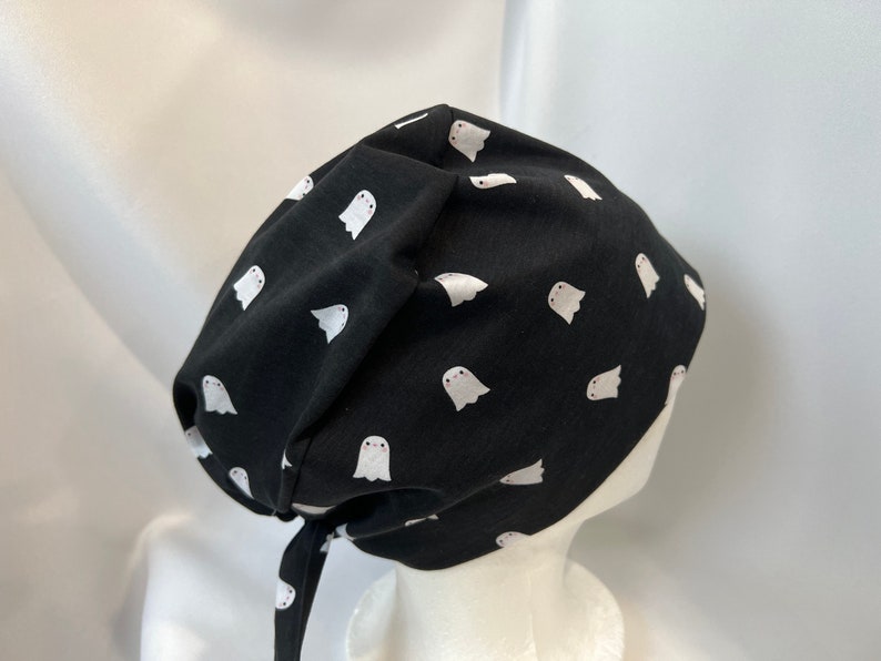 K&ouml;nnte beinhalten: Schwarze Stoffm&uuml;tze mit einem wei&szlig;en Geistermuster. Die M&uuml;tze ist hinten mit einem passenden Stoffband gebunden. Die M&uuml;tze ist zum Bedecken von Kopf und Haaren gedacht.