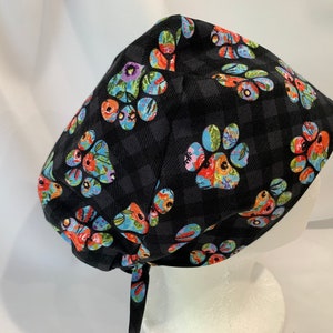 May include: Black scrub hat with a colourful paw print pattern. The hat has a tie at the back.