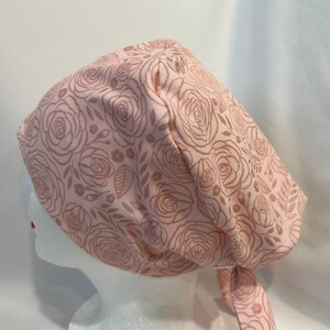 May include: A pink surgical scrub hat with a rose pattern. The hat has a tie at the back.