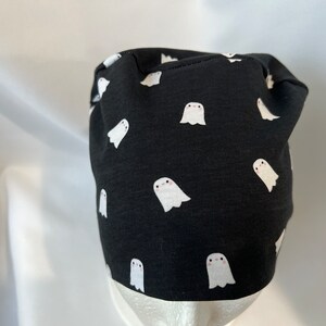 K&ouml;nnte beinhalten: Schwarze Beanie-M&uuml;tze mit einem sich wiederholenden Muster aus wei&szlig;en Cartoon-Geistern. Die M&uuml;tze ist aus einem dunklen Stoff gefertigt und eignet sich f&uuml;r Halloween oder den t&auml;glichen Gebrauch. Die Geister haben einfache, freundliche Gesichter.