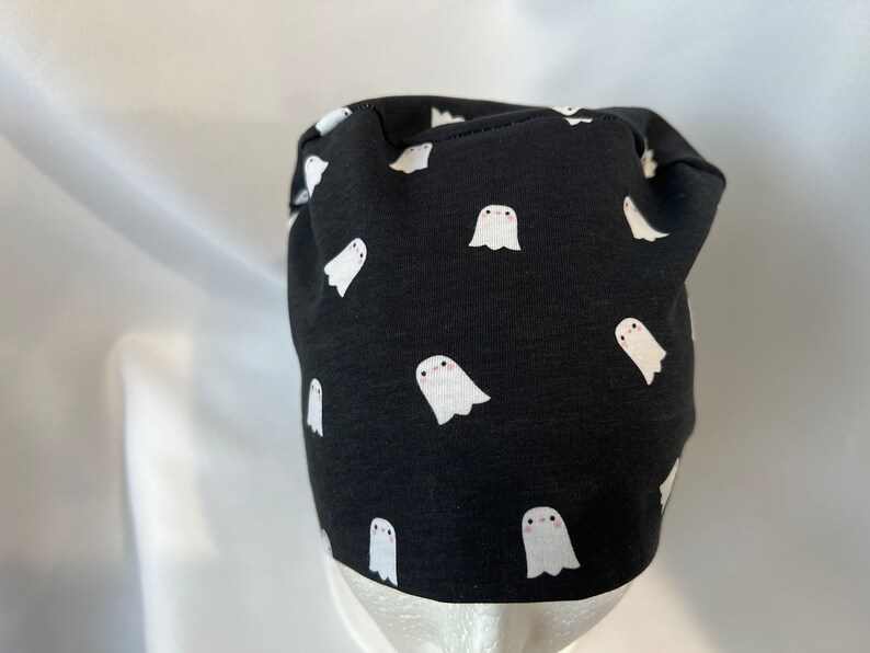 K&ouml;nnte beinhalten: Schwarze Beanie-M&uuml;tze mit einem sich wiederholenden Muster aus wei&szlig;en Cartoon-Geistern. Die M&uuml;tze ist aus einem dunklen Stoff gefertigt und eignet sich f&uuml;r Halloween oder den t&auml;glichen Gebrauch. Die Geister haben einfache, freundliche Gesichter.