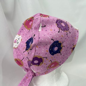 May include: Pink surgical scrub hat with a donut pattern. The donuts are purple, pink, and white with sprinkles and have cartoon faces.