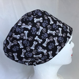 May include: Black scrub hat with a white and grey paw print and bone pattern. The hat has the word "bone" repeated throughout the pattern.