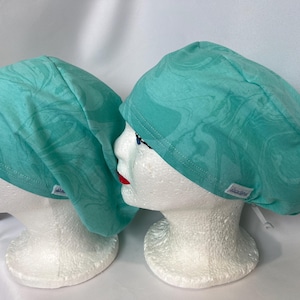 May include: Two turquoise blue scrub hats with a marbled pattern. The hats have a white label with the word "Scrubs" on them.
