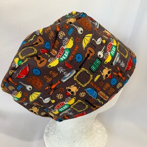 May include: A brown scrub hat with a colorful print of various icons from the TV show "Friends". The icons include a coffee cup, a guitar, a swan, a lobster, a key, a frame, and the words "Central Perk".