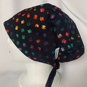 May include: Black scrub hat with a colourful paw print and heart pattern. The hat has a tie at the back.
