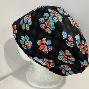 May include: A black scrub cap with a checkered design and vibrant paw prints. The paw prints are decorated with floral patterns in red, blue, and yellow. The cap is displayed on a white mannequin head.