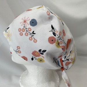 May include: White scrub hat with a floral print featuring pink, yellow, and blue flowers. The hat has a tie at the back.