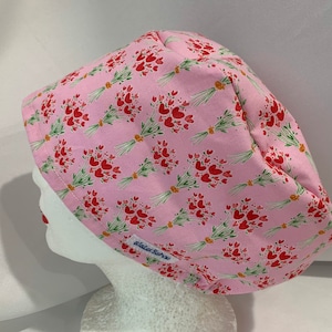 May include: Pink scrub hat with a floral pattern of red hearts and green stems. The hat has a blue label with the text "sister scrub"