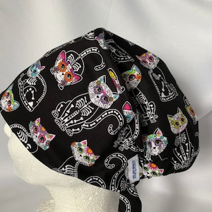 May include: Black scrub hat with a colorful Day of the Dead cat print. The cats have sugar skulls and floral designs. The hat has a tie at the back.