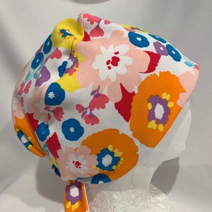 May include: A colorful floral scrub cap featuring large orange, pink, and blue flowers on a white background. The cap has a tie closure and is designed for medical professionals or those in the healthcare field.