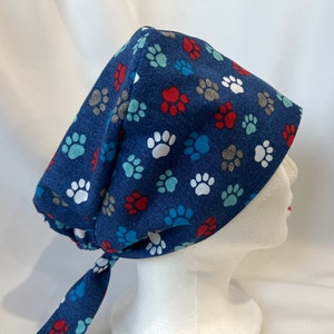 May include: A dark blue surgical scrub cap featuring a pattern of paw prints in white, red, blue, and brown. The cap is tied at the back and is designed to cover the head and hair.