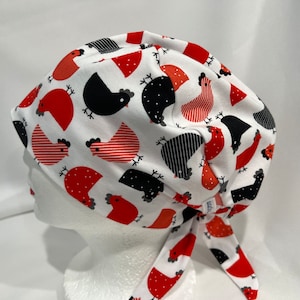 Puede incluir: Un gorro quirúrgico blanco con un estampado repetido de gallinas rojas y negras. Las gallinas tienen varios diseños, incluyendo patrones sólidos, rayados y punteados. El gorro está atado en la parte posterior con tela a juego.