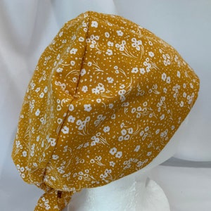 May include: Mustard yellow scrub cap with a white floral pattern. The cap is tied at the back and is made of a soft fabric. The cap is designed to be worn in a medical or professional setting.