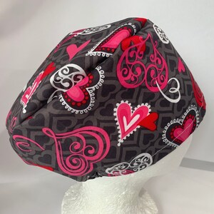 May include: A black scrub hat with a pink and white heart pattern. The hearts have a swirl design and are outlined in white.