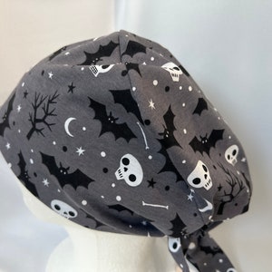 May include: Gray Halloween-themed scrub cap with a repeating pattern of black bats, skulls, stars, and bare trees. The cap is tied at the back and is made of a soft fabric.