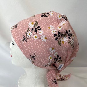May include: A pink surgical scrub hat with a white and black floral print and white polka dots. The hat has a tie at the back.