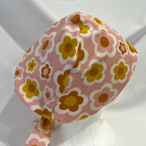 May include: A pink and brown floral print scrub hat with a tie. The hat has a large, bold floral pattern with white outlines.