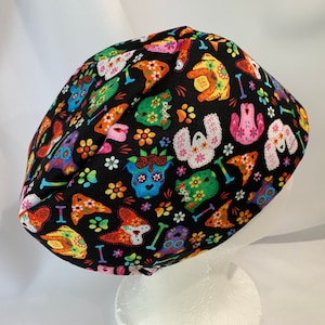 May include: A black surgical scrub cap featuring a colorful pattern of decorated dog skulls, flowers, and bones. The dog skull designs are in various colors, including orange, blue, pink, and green. The cap is made of fabric.