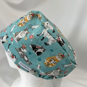 May include: A teal blue scrub hat with a white polka dot pattern and a repeating print of cats wearing flowers. The hat has a white label that says "elastic band".