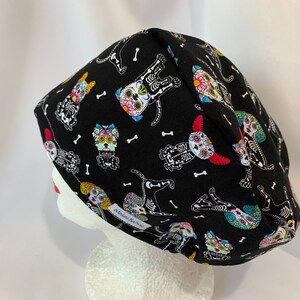 May include: Black scrub hat with a colorful Day of the Dead sugar skull dog pattern. The hat has a white label with the text "Scrubs & More" on it.