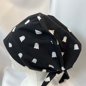 K&ouml;nnte beinhalten: Schwarze OP-Haube mit einem Muster aus wei&szlig;en Geistern. Die Haube wird hinten gebunden. Der Stoff ist ein weiches, leichtes Material, das f&uuml;r medizinische oder kulinarische Fachkr&auml;fte geeignet ist.