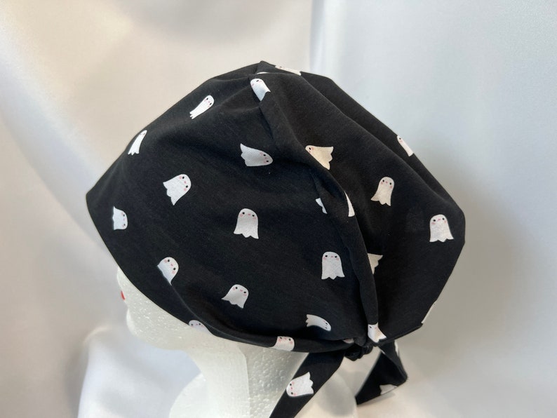 K&ouml;nnte beinhalten: Schwarze OP-Haube mit einem Muster aus wei&szlig;en Geistern. Die Haube wird hinten gebunden. Der Stoff ist ein weiches, leichtes Material, das f&uuml;r medizinische oder kulinarische Fachkr&auml;fte geeignet ist.