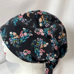 May include: A black scrub hat with a floral print featuring pink, blue, and green flowers on a black background. The hat has a tie that can be tied at the back of the neck.