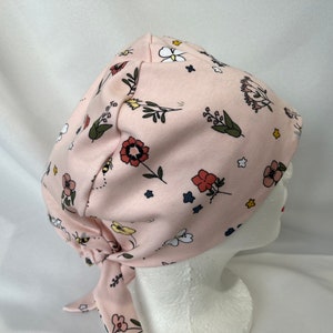 May include: A pink scrub hat with a floral print featuring bees, flowers, and stars. The hat has a tie at the back.
