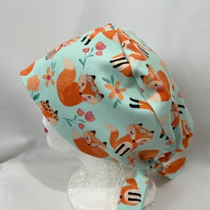 May include: A turquoise blue scrub hat with a tie back. The hat features a repeating pattern of orange foxes with floral accents.