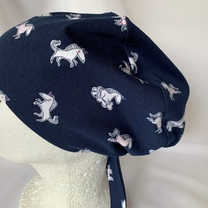 May include: A navy blue scrub hat with a white unicorn print. The hat has a tie that can be tied at the back of the neck.