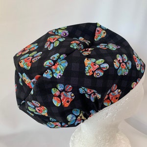 May include: Black scrub hat with a floral paw print pattern. The hat is made of a soft, comfortable fabric and has a tie at the back.