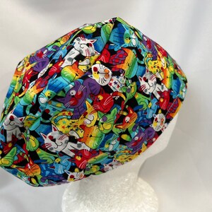 May include: A black scrub hat with a colourful print of cartoon cats in various poses. The cats are in shades of red, blue, green, purple, and white.