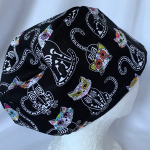 May include: Black scrub hat with a colorful Day of the Dead cat pattern. The cats are skeletonized and have floral details on their faces.