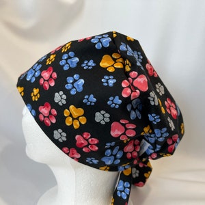 May include: Black scrub hat with a colourful paw print pattern. The hat has a tie at the back.