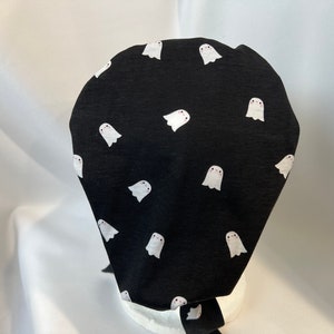 K&ouml;nnte beinhalten: Schwarze Stoffm&uuml;tze mit einem sich wiederholenden Muster aus wei&szlig;en Cartoon-Geistern. Jeder Geist hat eine kleine rosa R&ouml;te und einfache Augen. Die M&uuml;tze ist hinten gebunden. Die M&uuml;tze ist wahrscheinlich f&uuml;r medizinische oder kulinarische Zwecke bestimmt.