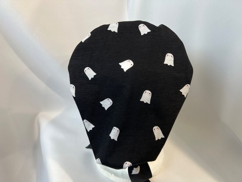 K&ouml;nnte beinhalten: Schwarze Stoffm&uuml;tze mit einem sich wiederholenden Muster aus wei&szlig;en Cartoon-Geistern. Jeder Geist hat eine kleine rosa R&ouml;te und einfache Augen. Die M&uuml;tze ist hinten gebunden. Die M&uuml;tze ist wahrscheinlich f&uuml;r medizinische oder kulinarische Zwecke bestimmt.