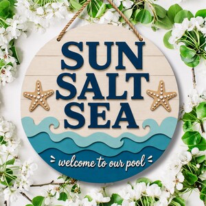 以下が含まれることがあります： SUN SALT SEAという濃い青色の文字と、welcome to our poolという白色の文字が書かれた円形の木製看板。デザインには2つのベージュ色のヒトデと、下部に重なり合うティール色と濃い青色の波が描かれており、茶色のロープで吊り下げられている。