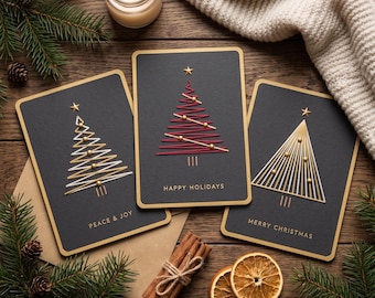 Può includere: Tre biglietti d'auguri rettangolari neri con bordi dorati. Ogni biglietto presenta un design stilizzato di albero di Natale composto da linee sottili e piccole sfere dorate, sormontato da una stella. Il testo recita PEACE & JOY, HAPPY HOLIDAYS e MERRY CHRISTMAS.